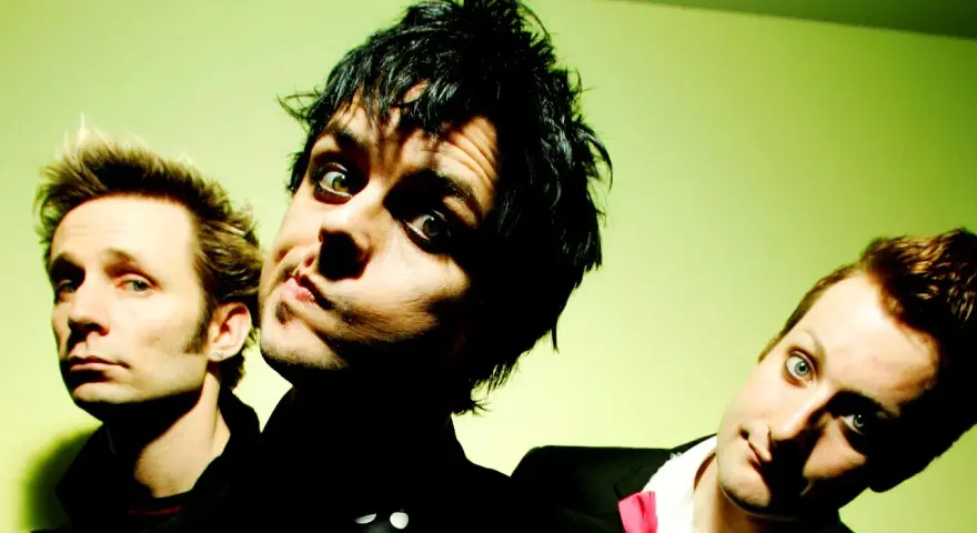Trzej członkowie Green Day: Billie Joe Armstrong, Mike Dirnt i Tré Cool, pozują do zdjęcia na zielonym tle.