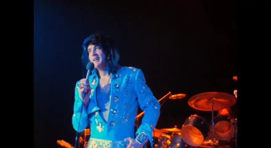 Elvis Presley, in einem glitzernden blauen Anzug, singt ins Mikrofon. Ein Bild, das an seine legendären Elvis Presley Filme erinnert.