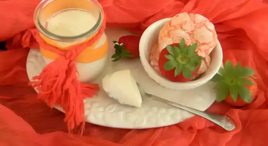 Yogur cremoso en frasco con fresas y trozos de yogur en un plato blanco sobre tela naranja.