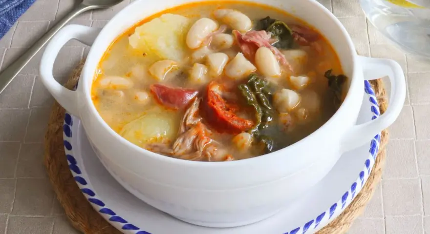 Un tazón blanco de caldo gallego con patatas, alubias, chorizo y grelos.
