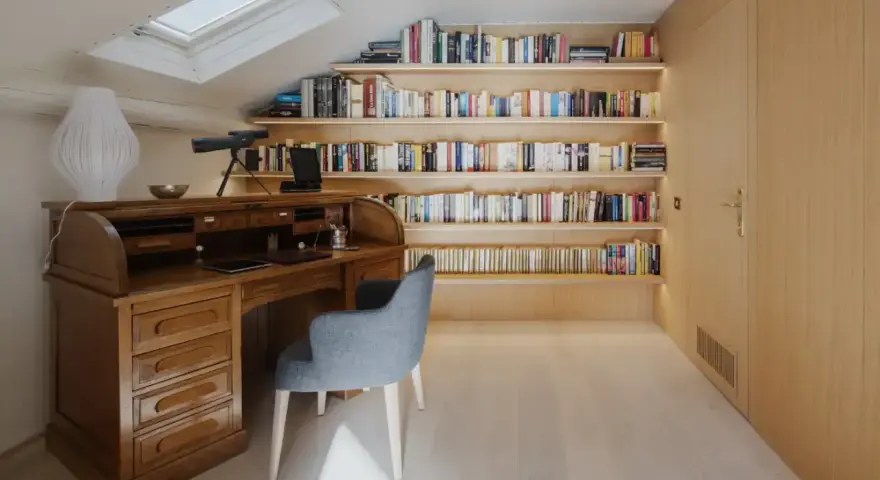 Scrivania in legno massiccio e libreria moderna illuminata, un perfetto esempio di come abbinare e mischiare arredamento classico e moderno in uno studio mansardato.
