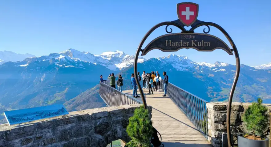 Tourists enjoy the breathtaking panorama from the Harder Kulm viewpoint, with snow-capped mountains under a clear blue sky.