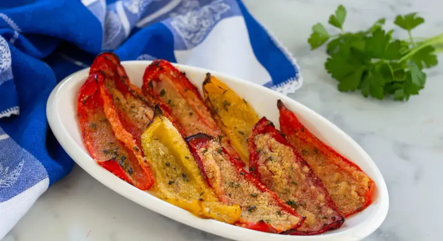 Peperoni gratinati al forno, rossi e gialli, cotti alla perfezione con pangrattato e erbe aromatiche, serviti su un piatto bianco con un tovagliolo blu.