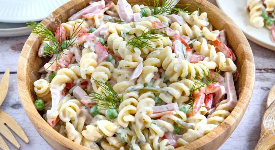 Ein großer Holzteller voller cremigen Nudelsalat mit Schinken, Erbsen und Paprika. Mit Dill garniert, sieht er aus wie der beste Nudelsalat!