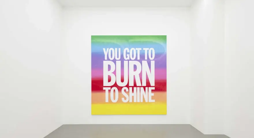 Œuvre de John Giorno dans une galerie, avec le message "YOU GOT TO BURN TO SHINE" sur un fond arc-en-ciel.