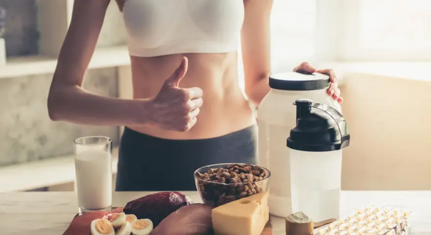Kobieta z kciukiem w górę prezentuje produkty wspierające dietę wysokobiałkową: mięso, rybę, jajka, ser, orzechy, mleko i suplementy.