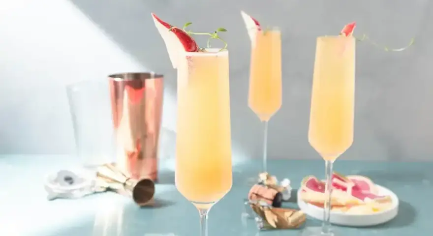Tre eleganta glas med en ljusgul drink med gin och prosecco, garnerade med en tunn skiva röd frukt och en kvist.