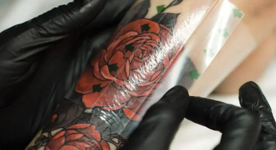 Ręce w czarnych rękawiczkach zdejmują folię ochronną z tatuażu z czerwonymi różami. To kluczowy etap, jak zdjąć second skina, by tatuaż prawidłowo się goił.