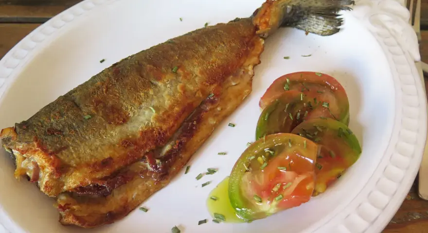 Trucha a la navarra dorada y crujiente, servida con rodajas de tomate fresco y hierbas aromáticas.