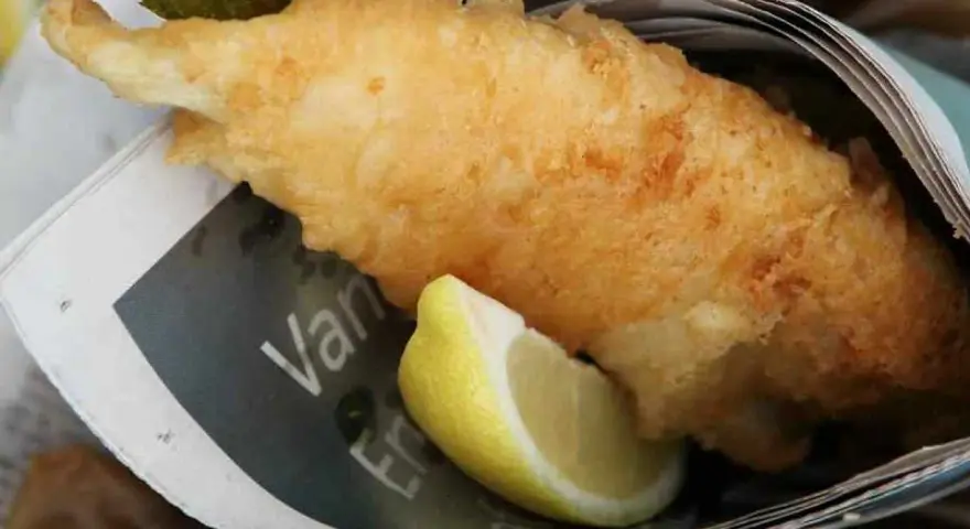Krispigt friterad fisk med citronklyfta, inslagen i tidningspapper. En klassisk fish and chips-upplevelse.
