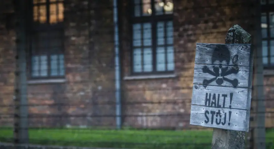 Tablica z symbolem czaszki i piszczeli oraz napisem "HALT! STÓJ!" na tle ceglanego budynku i drutu kolczastego, przypominająca o grozie obozów koncentracyjnych.