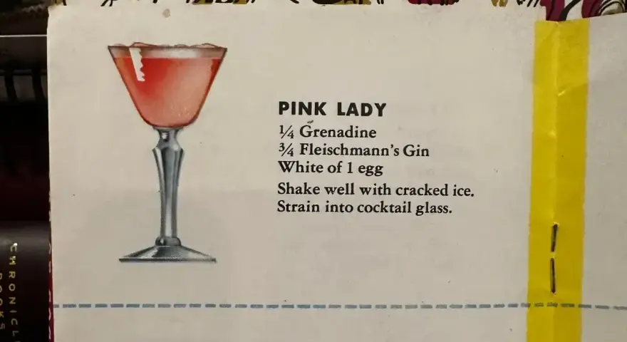 Elegancki kieliszek z różowym drinkiem Pink Lady, obok przepis na jego przygotowanie: grenadyna, gin i białko jaja.