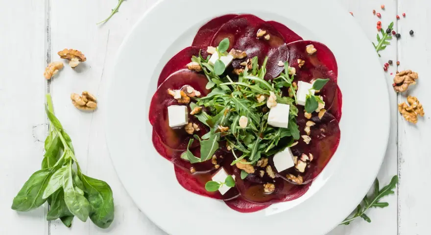 Eleganckie carpaccio z buraka z rukolą, orzechami włoskimi i kostkami sera feta na białym talerzu.