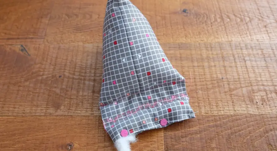 Un berlingot tissu gris à petits cœurs roses et blancs, en cours de rembourrage, est posé sur un parquet en bois.