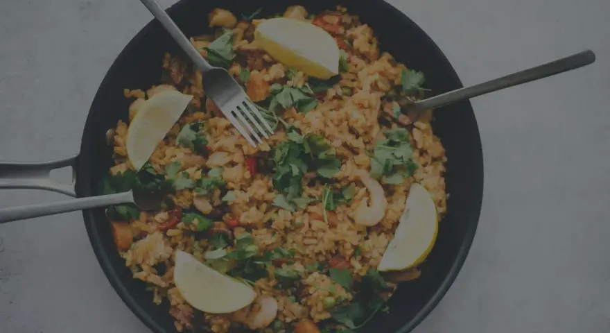 En stor panna med jollof rice, räkor, citronklyftor och färsk persilja. Tre bestick ligger redo att avnjutas.