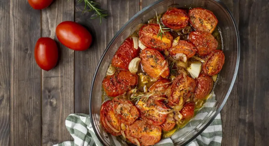 Pomodori gratinati al forno, dorati e succosi, con cipolle e rosmarino in una teglia di vetro su un tavolo di legno.