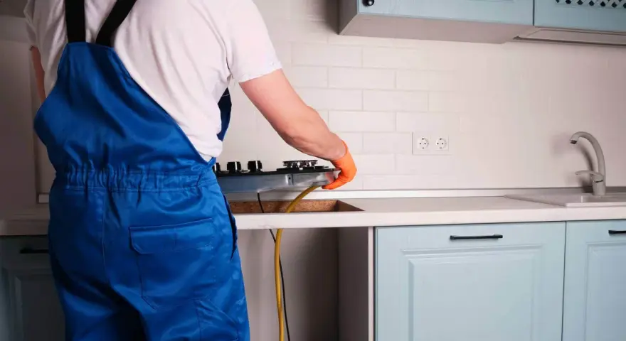 Instalator w niebieskim kombinezonie podłącza kuchenkę gazową w kuchni.