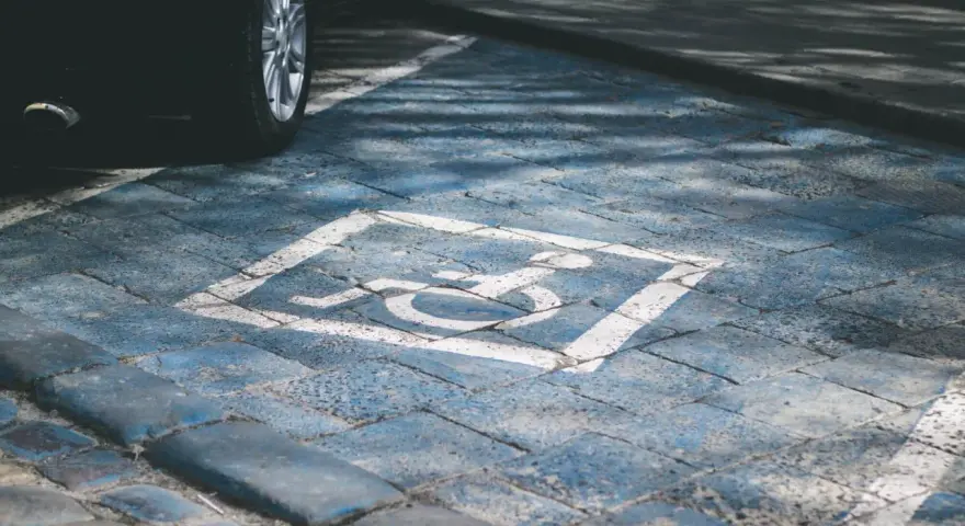 Niebieskie miejsce parkingowe dla niepełnosprawnych pod blokiem, z symbolem wózka inwalidzkiego. Obok widoczna część czarnego samochodu.