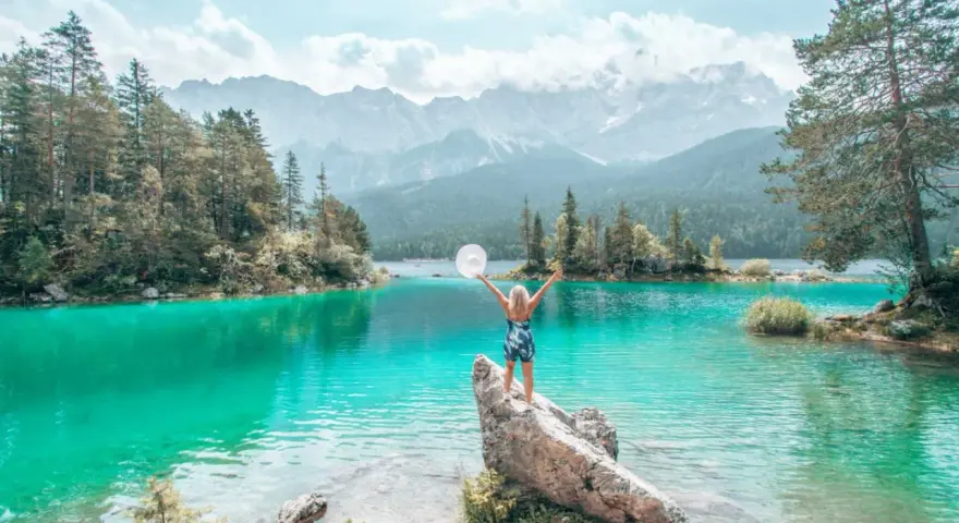 Frau mit ausgebreiteten Armen und Hut am türkisfarbenen Eibsee in Bayern, im Hintergrund Berge und Wald.
