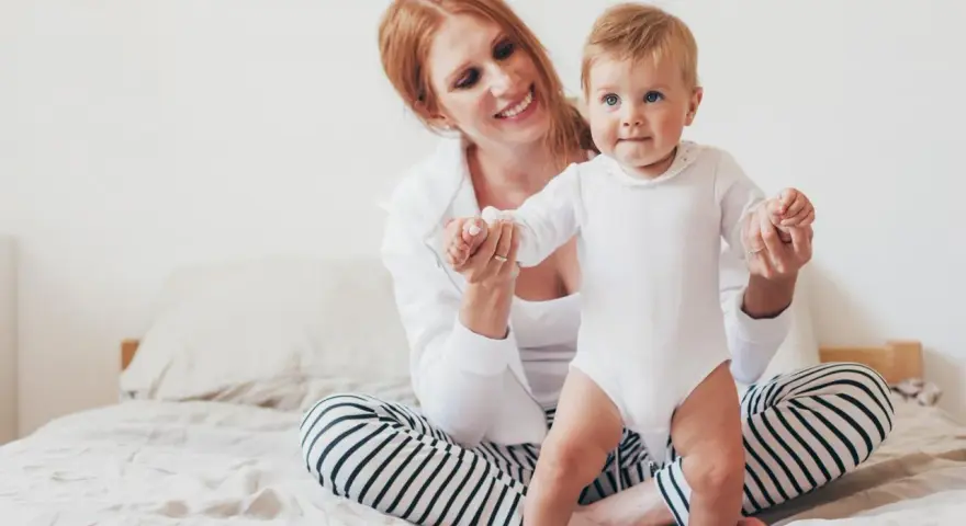 Uśmiechnięta mama pomaga swojemu 8 miesięcznemu dziecku w pierwszych krokach na łóżku.