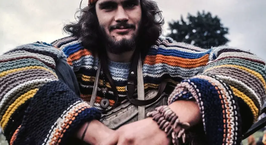 Młody mężczyzna w kolorowym swetrze w paski i opasce na głowie, uśmiecha się. Wygląda jak prawdziwy hippie.