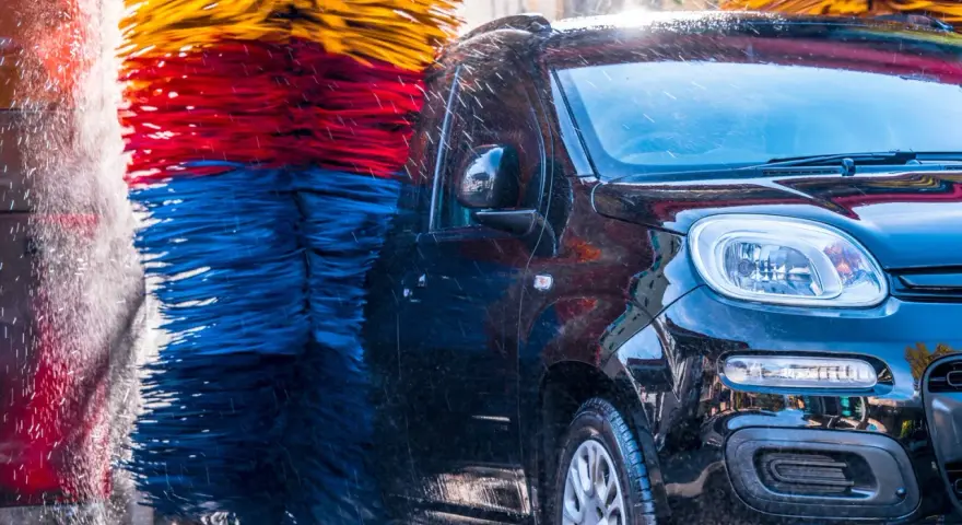 Czarna, błyszcząca karoseria samochodu w myjni automatycznej, otoczona kolorowymi szczotkami. Czy myjnia automatyczna niszczy lakier? Woda spływa po aucie.