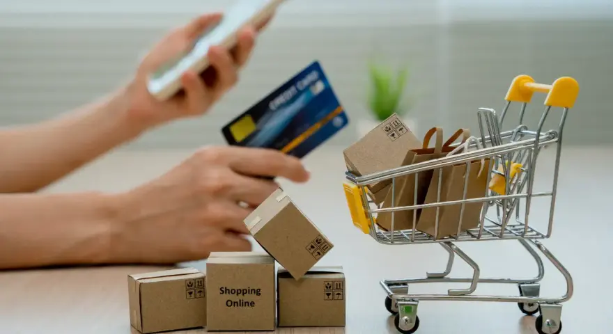 Manos sostienen un teléfono y tarjeta de crédito, junto a un carrito de compras con cajas y bolsas. El modelo 369 representa la facilidad de comprar en línea.