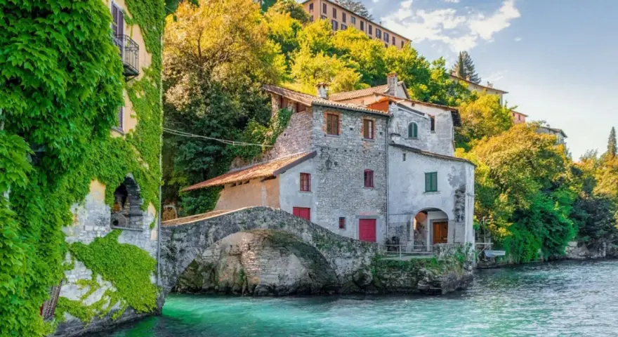 Antica casa in pietra con edera su un lago turchese, un ponte ad arco e colline verdi. Un luogo incantevole per i cammini in Lombardia.