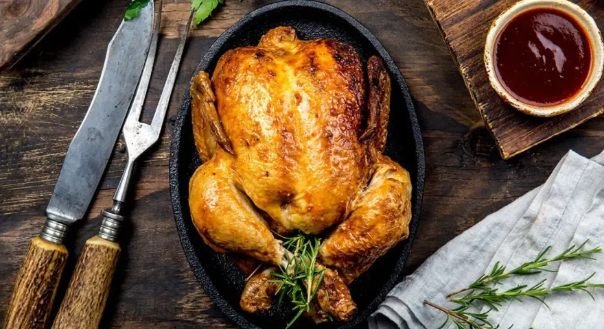 Pollo al horno dorado y jugoso, servido en sartén de hierro fundido con romero. Acompañado de salsa barbacoa y cubiertos rústicos.