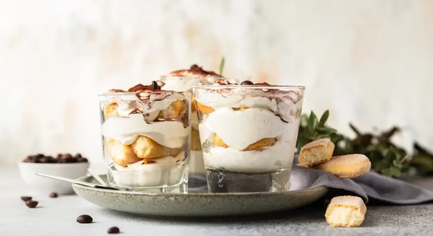 Pyszne desery włoskie: tiramisu w szklankach, udekorowane kakao i kawą. Obok kawałki biszkoptów i ziarna kawy.