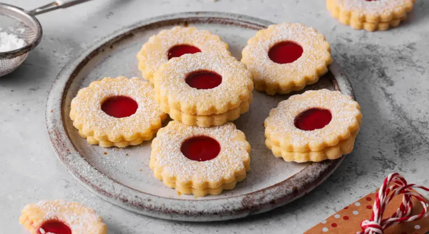 Ein Teller voller **die besten Spitzbuben der Welt**, bestäubt mit Puderzucker, mit leuchtend roter Marmeladenfüllung.