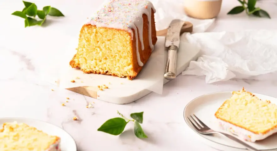 Saftiger zitronenkuchen kastenform mit Zuckerguss und bunten Streuseln, auf einem Marmorsockel, daneben ein Messer und ein Teller mit einem Stück Kuchen.