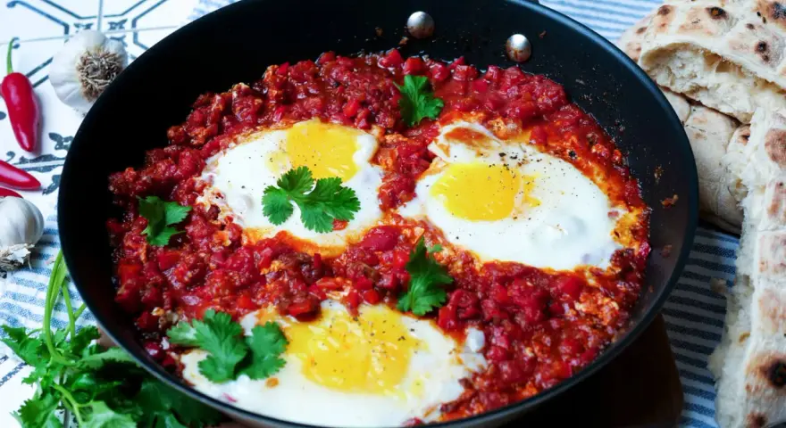 Herzhaftes Shakshuka-Rezept in einer Pfanne mit pochierten Eiern in würziger Tomatensoße, garniert mit Koriander. Dazu gibt es Fladenbrot.