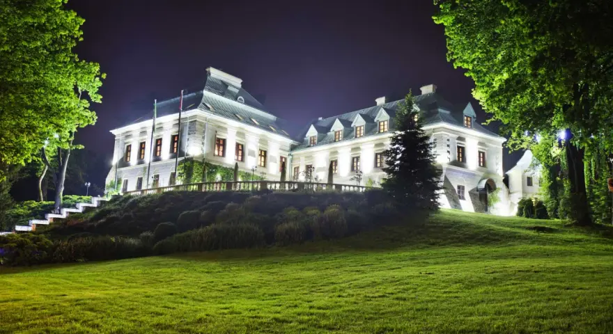 Elegancki hotel w lesie nocą, oświetlony blaskiem okien i zewnętrznych lamp. Otoczony zielenią, zaprasza do odpoczynku.
