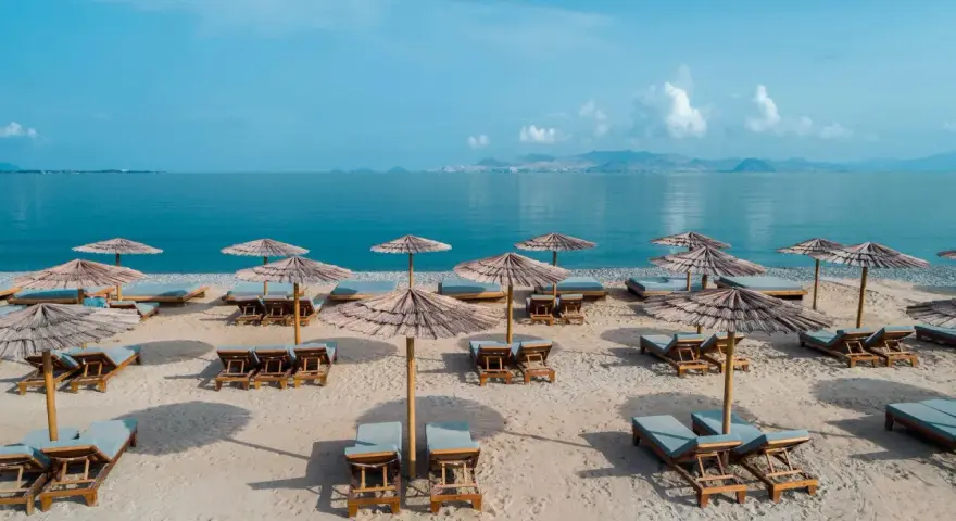 Plaża z leżakami i parasolami, idealna na relaks nad morzem. W oddali widać góry i błękitne niebo.