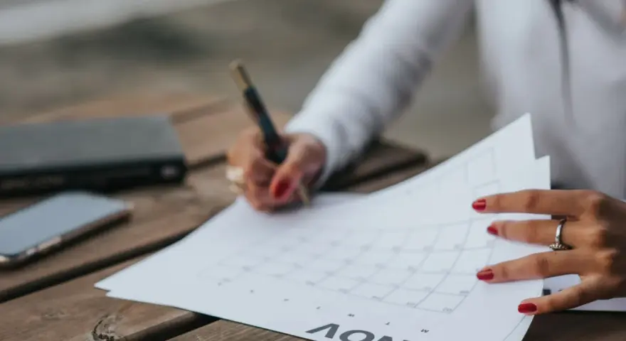 Une main avec des ongles rouges écrit sur un calendrier, planifiant peut-être la date de mariage. Un téléphone et un carnet sont à proximité.