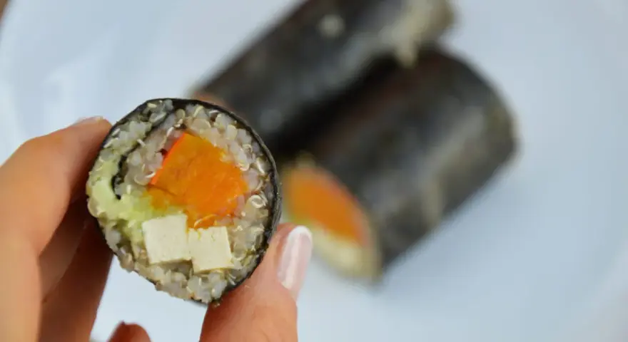 Rolka sushi z tofu, dynią i awokado, trzymana w dłoni.