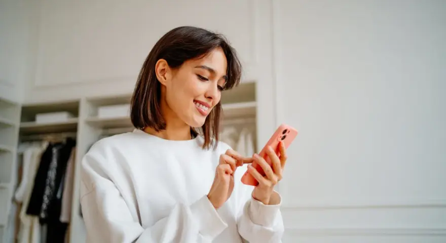 Uśmiechnięta kobieta przegląda telefon w swojej garderobie. Jej gotowy biogram czeka na publikację.