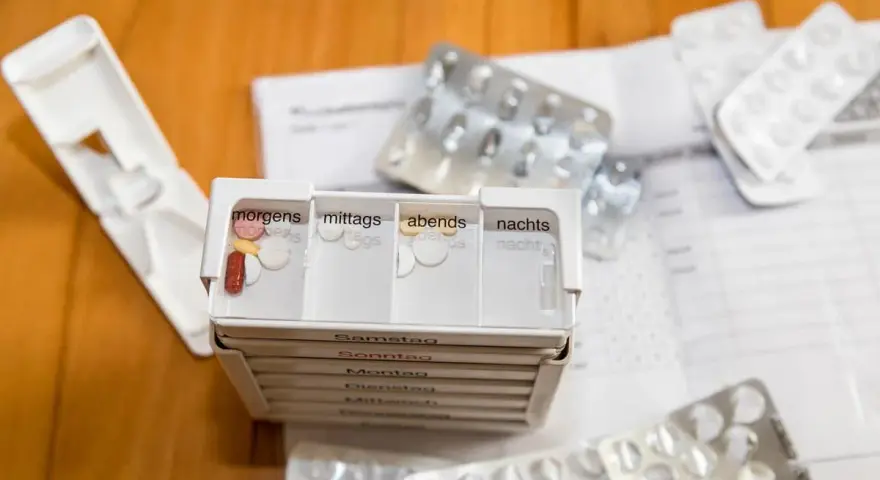 Tablettendose mit Fächern für morgens, mittags, abends, nachts. Leere Blister und ein Medikament liegen daneben.