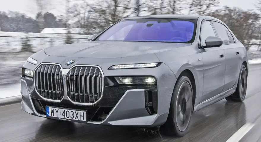 Szary BMW M7 pędzi po mokrej drodze, otoczony zimowym krajobrazem.