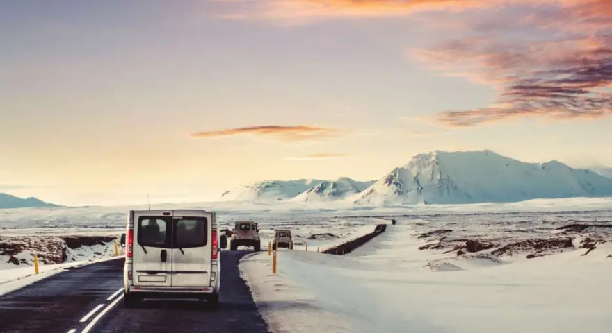 A silver van leads a convoy of vehicles driving in Iceland in December, past snow-covered mountains under a dramatic sunset sky.