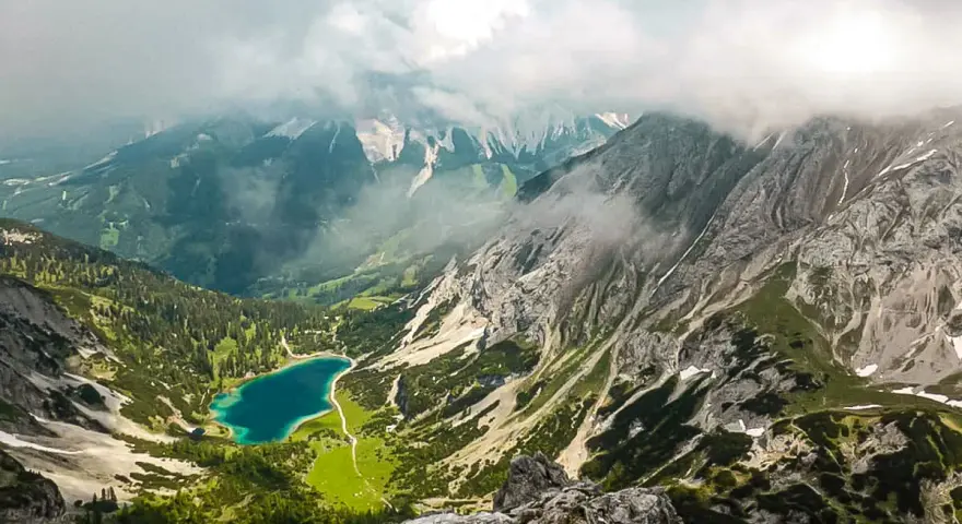 Blick vom Drachenkopf auf einen türkisfarbenen Bergsee, umgeben von grünen Hängen und felsigen Gipfeln, Wolken ziehen über die Landschaft.