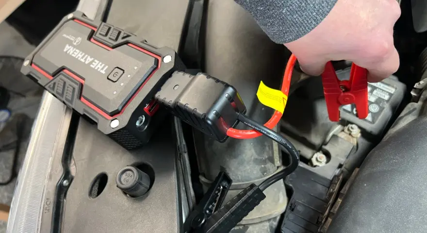 A hand connects red jumper cables from "The Athena" lithium jump starter to a car battery, demonstrating how lithium jump starters work by providing a temporary power boost.