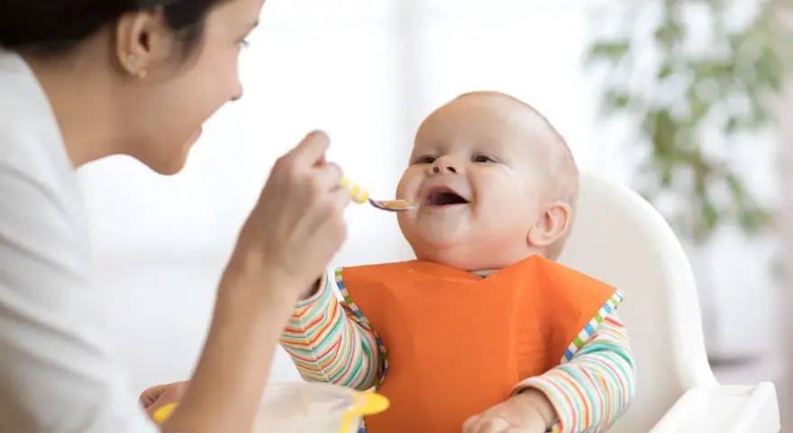 Mama karmi uśmiechniętego niemowlaka łyżeczką. To idealny przykład, co na śniadanie dla niemowlaka – zdrowo i radośnie!