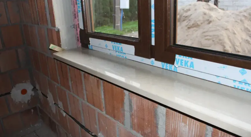 Montaż parapetów zewnętrznych po ociepleniu. Nowy, jasny parapet lśni pod brązową ramą okna, a ceglana ściana czeka na dalsze prace.