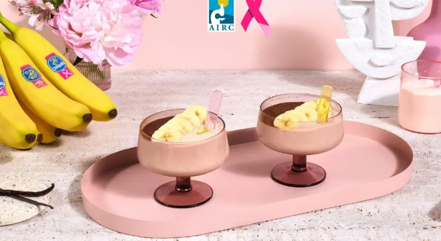 Due coppe di cremoso gelato alla banana, guarnite con fettine di banana e cacao, su un vassoio rosa. Banane fresche e fiori rosa sullo sfondo.