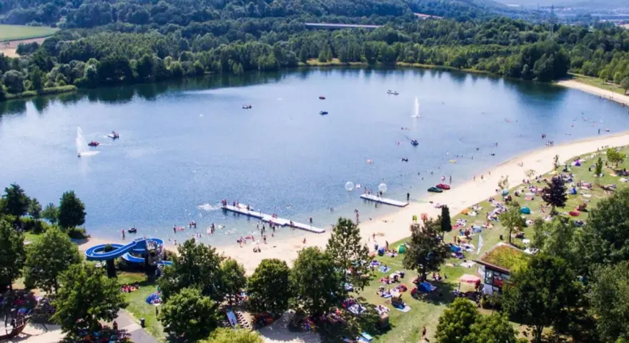 Sommerliche Idylle am Borkener See mit vielen Badegästen, Wasserrutsche und schattigen Plätzen unter Bäumen.
