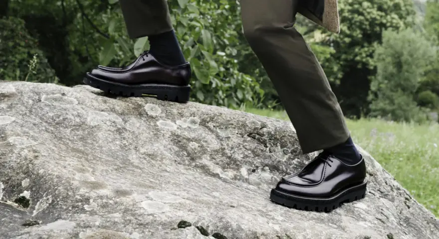 Scarpe oxford derby lucide su una roccia, con pantaloni verdi e sfondo verdeggiante.