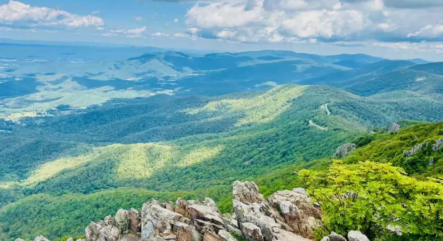 Scenic mountain vista, perfect for day trips from Richmond VA. A winding road snakes through lush green hills under a bright, cloudy sky.