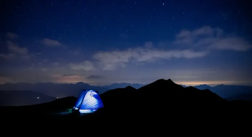 Une tente bleue s'illumine sous un ciel étoilé, témoin silencieux d'un trek inoubliable au cœur des montagnes.